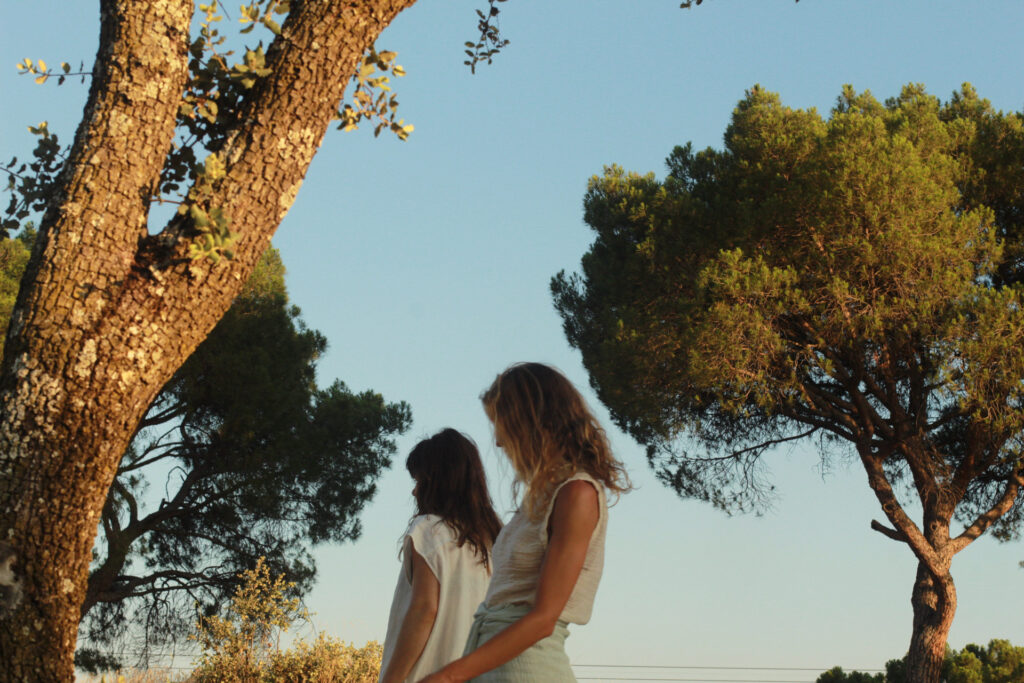 Back view of two women walking outdoors among tall pine trees at sunset, with warm natural light and a clear sky
